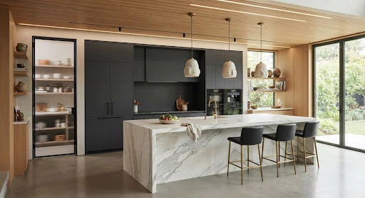 A high-end architectural kitchen with a monumental 12-foot waterfall quartzite island, matte charcoal handleless slab cabinets, integrated induction cooktop, fluted glass walk-in scullery pantry, brushed brass architectural faucet, warm wood ceiling slats and seamless architectural lighting.