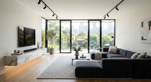 Sleek contemporary living room with floating marble media console, matte black track lights, low profile charcoal velvet modular sectional, and floor-to-ceiling glass fenestrations.